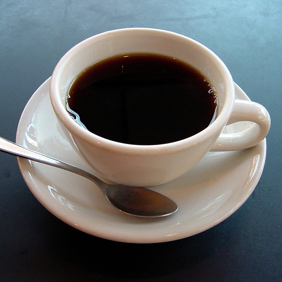 Image of a white ceramic coffee cup on a saucer plate with a silver spoon on top of a blue surface.