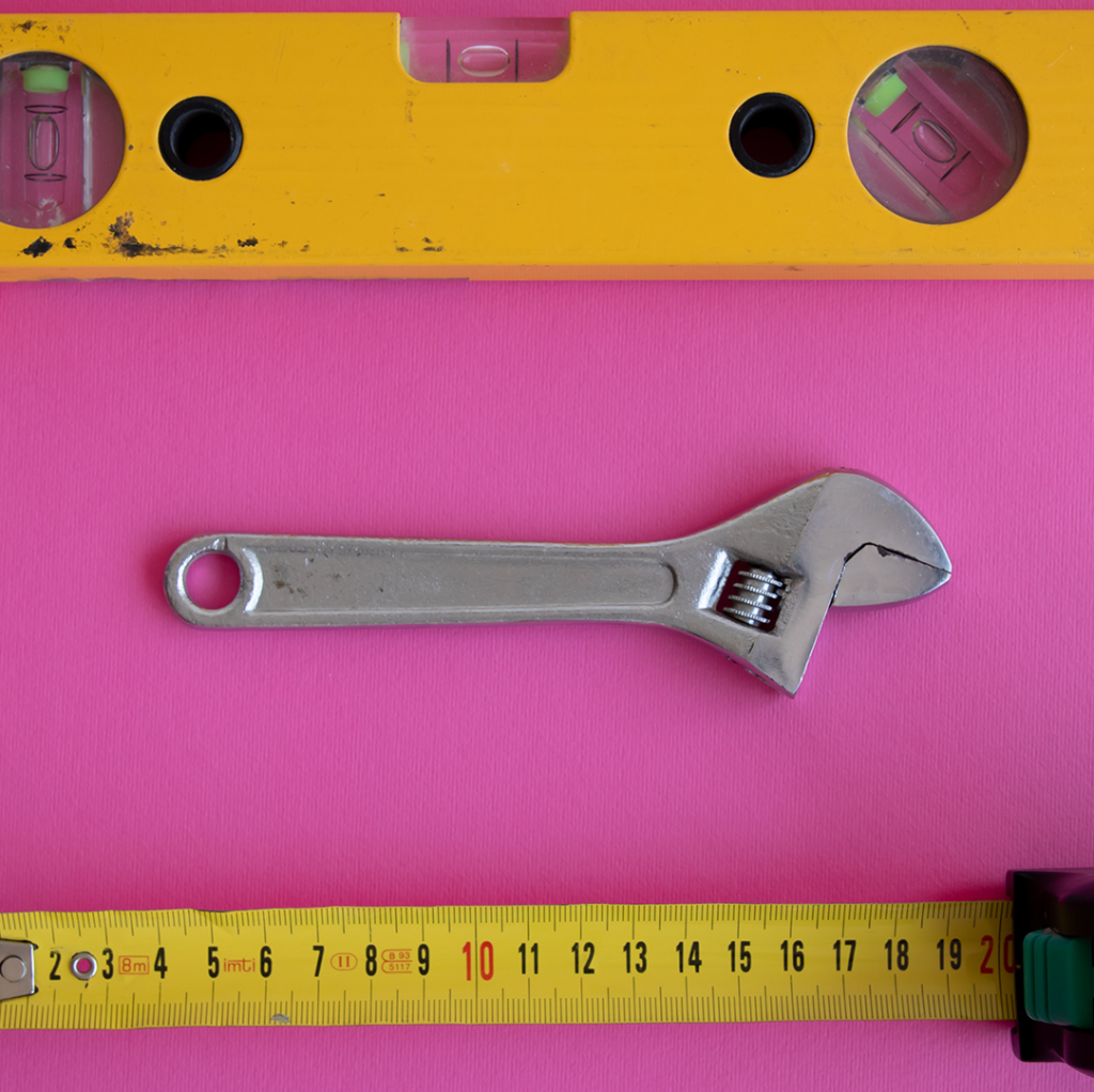 Photo of a yellow level tool, a wrench, and an extended tape measure on a pink surface.
