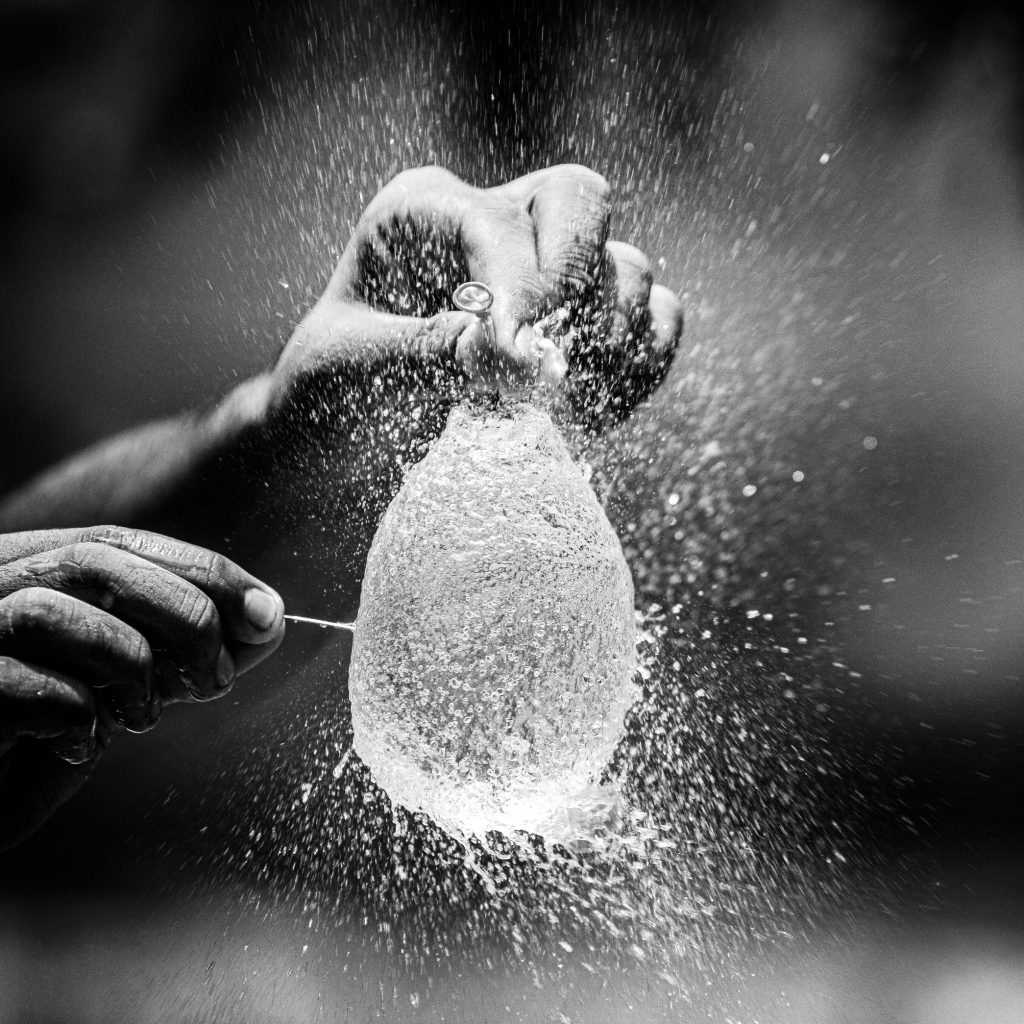 Black and white photo of a hand holding a water ballon as it is being popped by a pin.