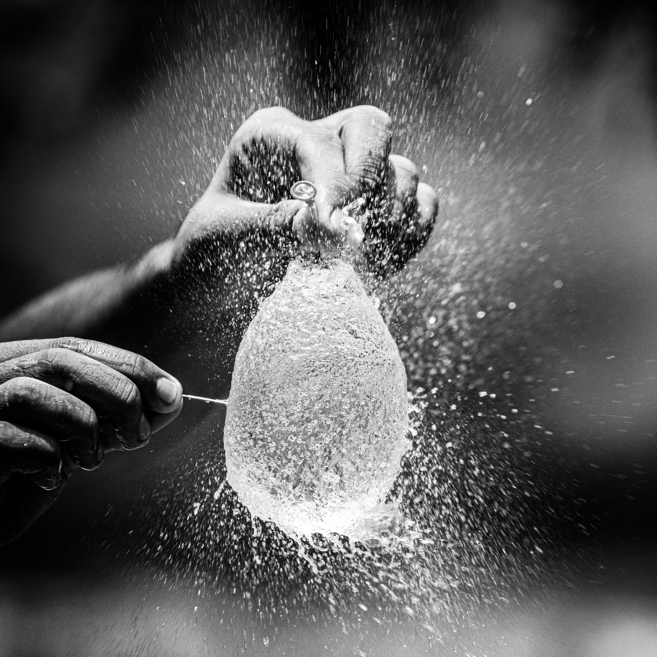 Black and white photo of a hand holding a water ballon as it is being popped by a pin.