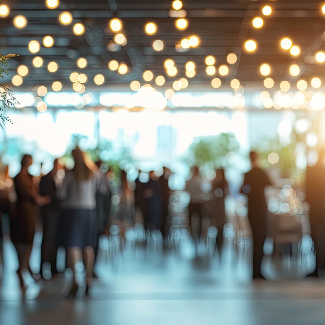 Blurred image of people mingling at a social event under warm lights.