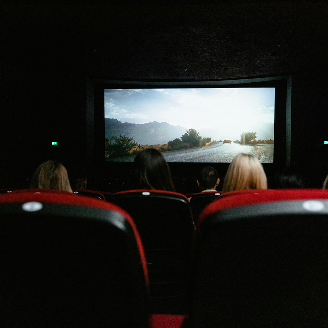 Photo from the back of a movie theater with a handful of people sitting in red upholstered chairs in front of movie screen playing.